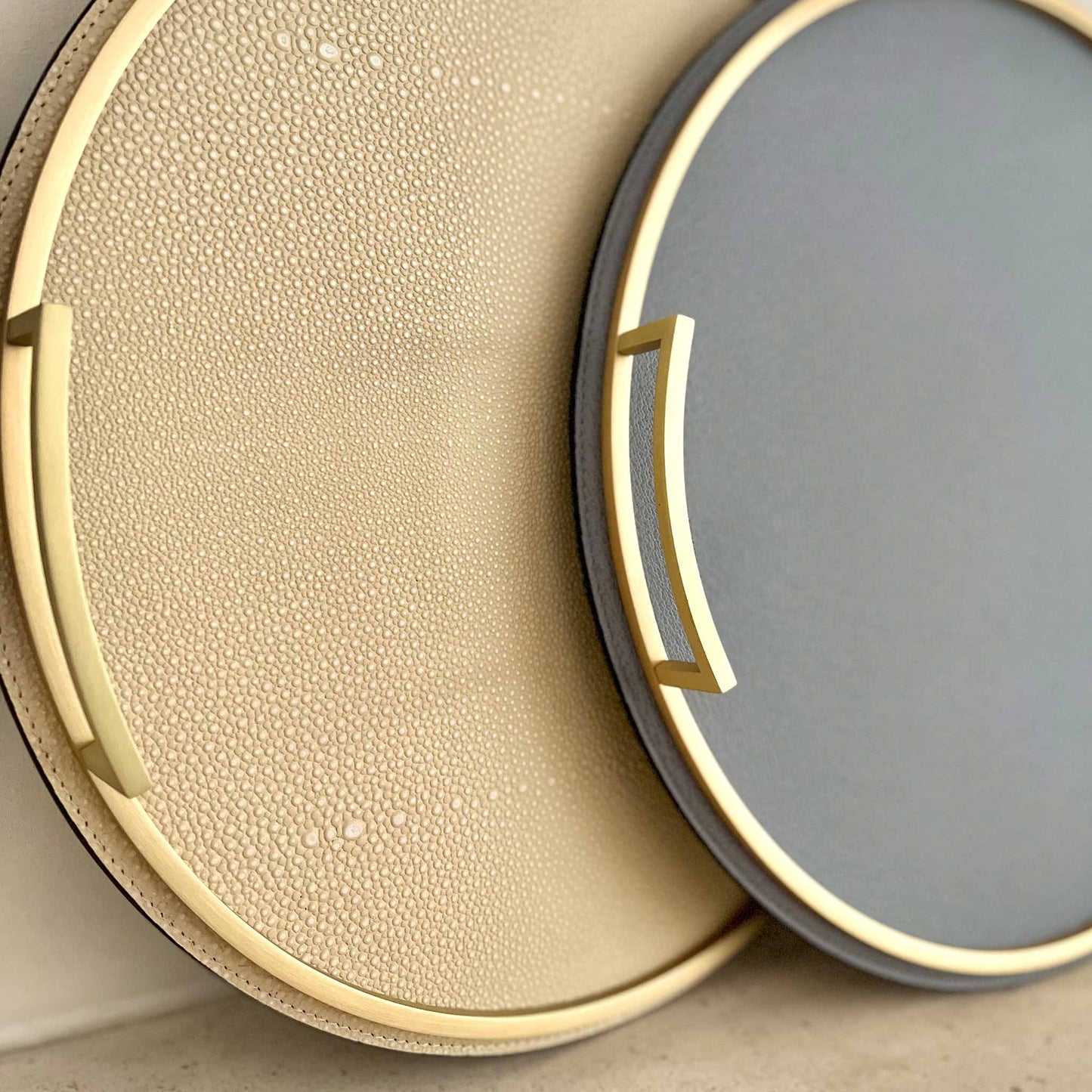 Close up of grey-blue round leather tray with cream tray with shagreen texture, both with brass handles