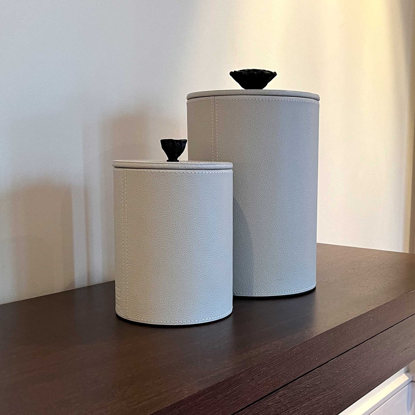 Lifestyle image of matching Close up lifestyle image of two light cream leather cylinder boxes with cast brass handles. Displayed on a luxury console