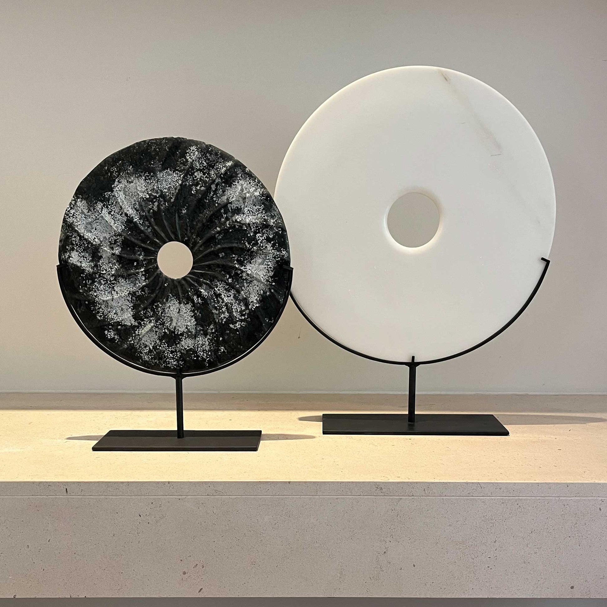 Lifestyle image of distressed black marble bi disc with shell type grooves displayed on a bronze stand. Next to larger plain white marble disc, also on a marble stand.