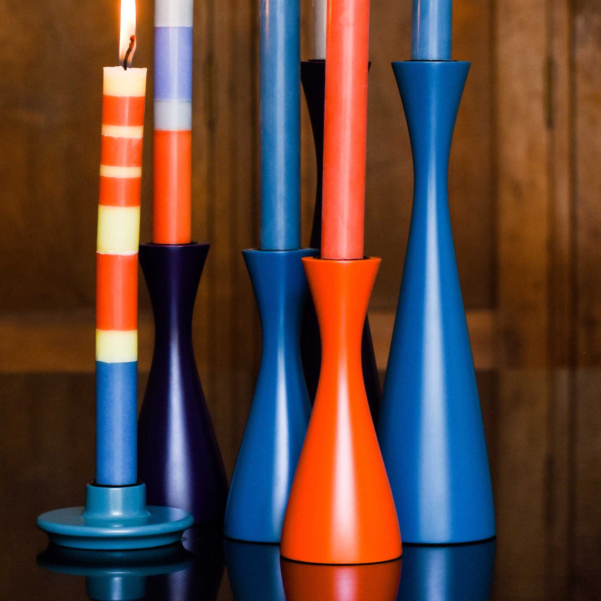 Close up of lifestyle image of several blue and red candleholders in a variety of heights with striped tall candles. Displayed against a wood panelled background.