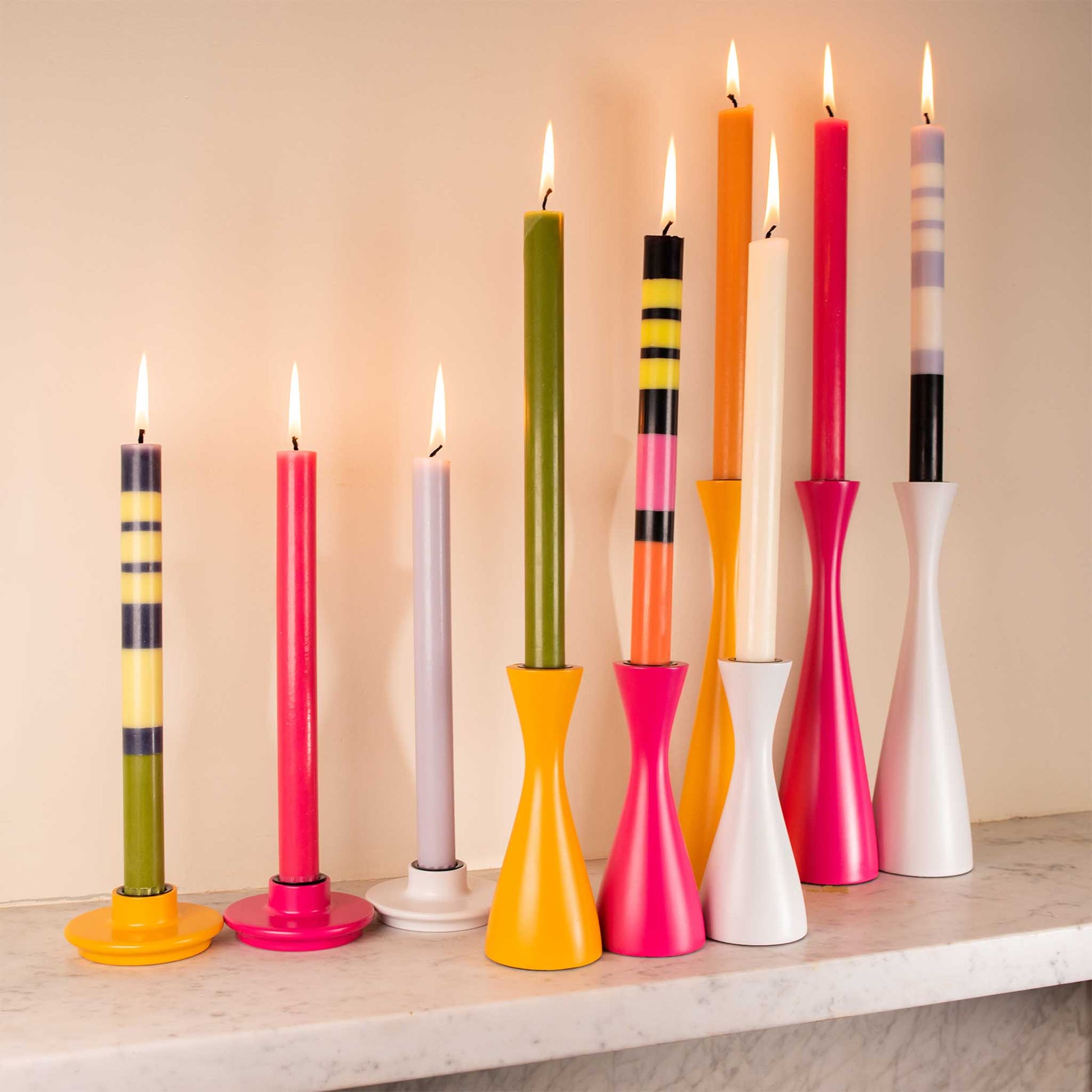 Lifestyle image of minimal brightly multi coloured candleholder with a variety of tall candles displayed on a mantelpiece