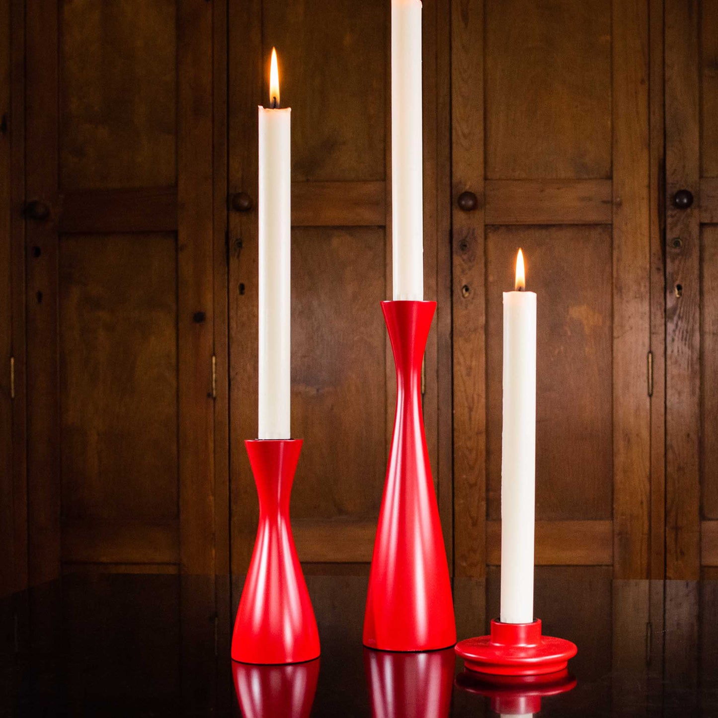 Lifestyle image of red candleholders in a variety of heights with white tall candles. Displayed against a wood panelled background.