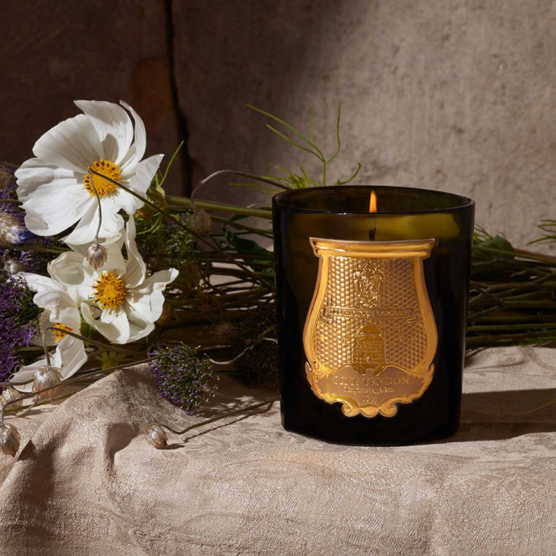 lifestyle image of a lit green jar cire trudon candle next to some flowers