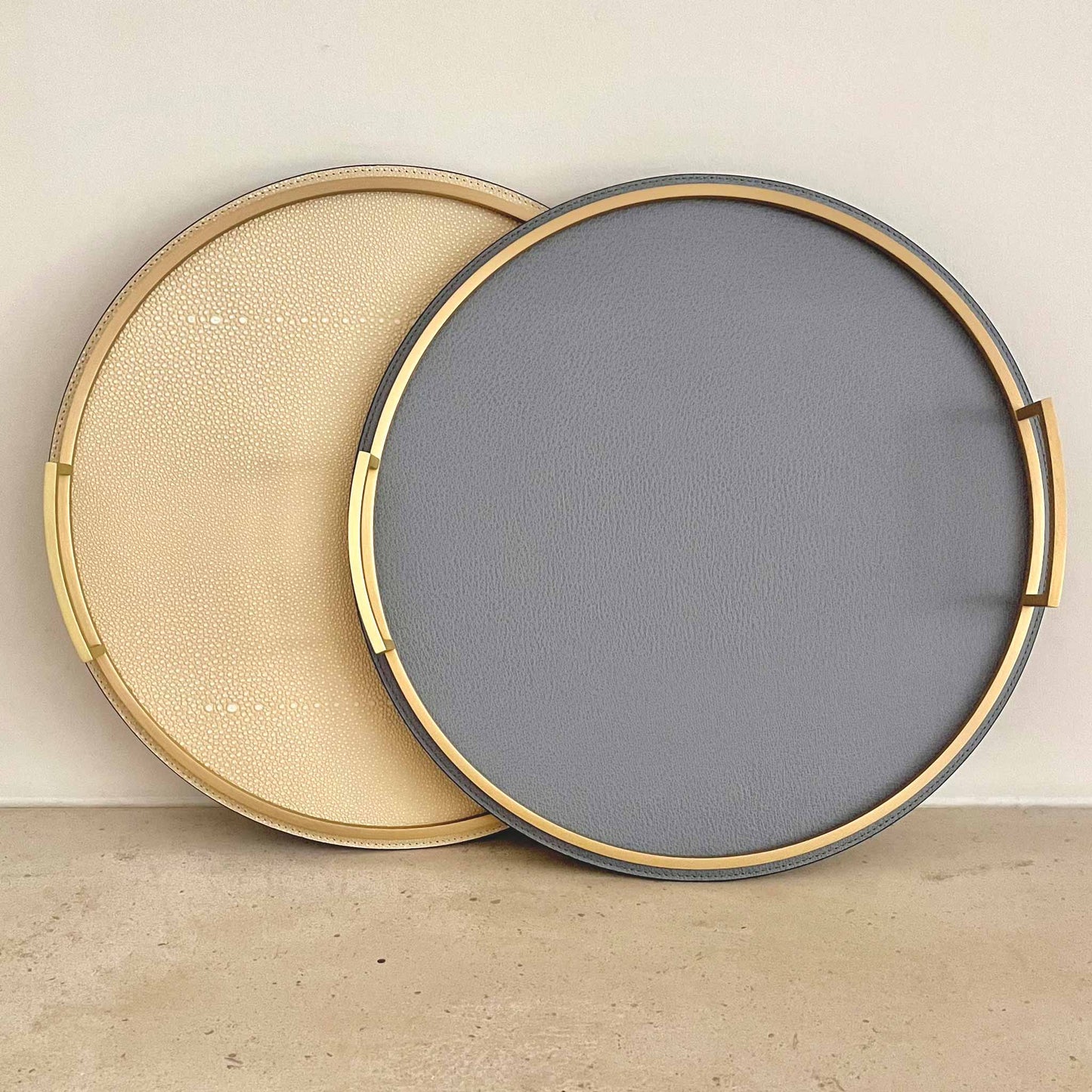 Two round textured Italian leather trays with brass handles. One cream coloured and another storm blue. 