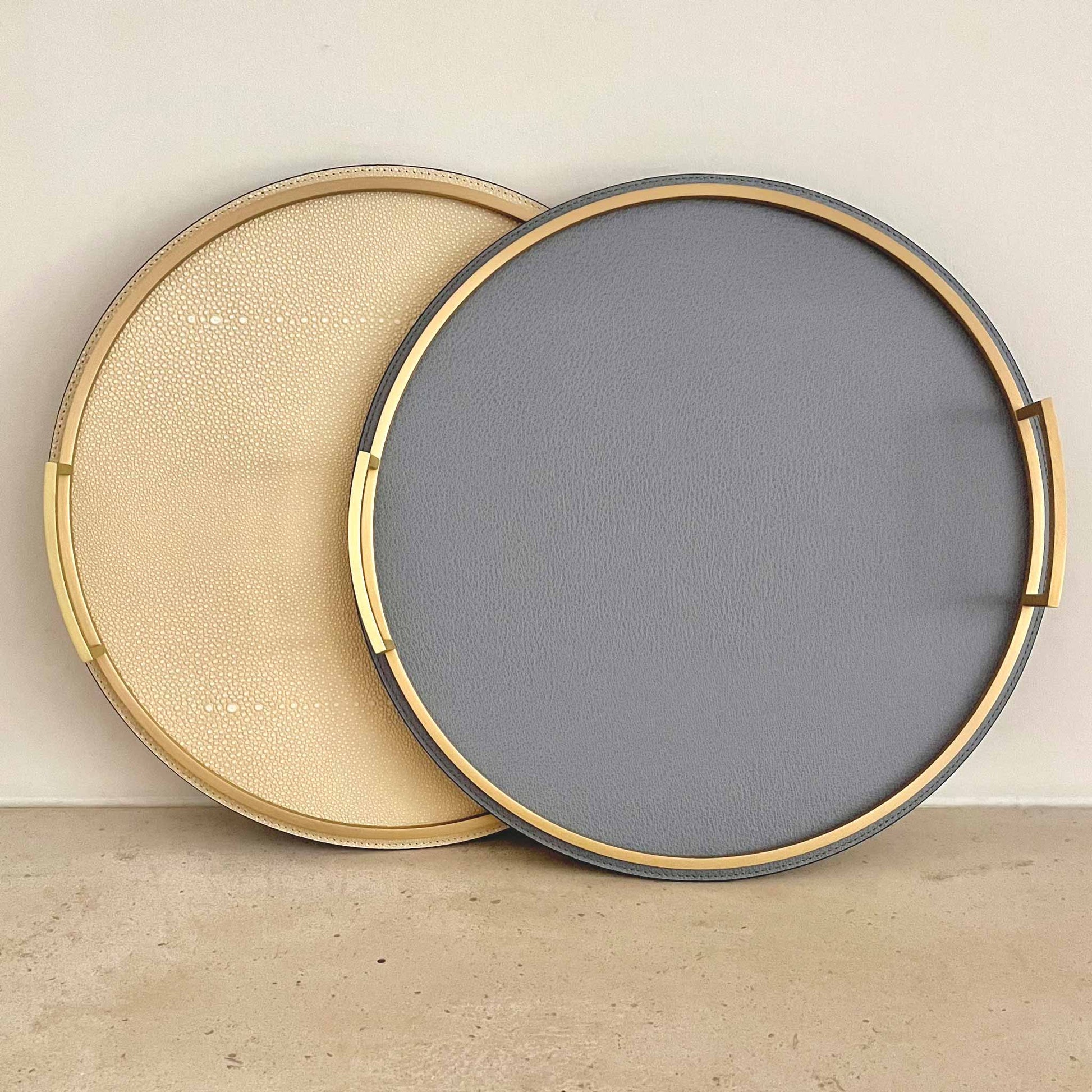 Two round textured Italian leather trays with brass handles. One cream coloured and another storm blue. 