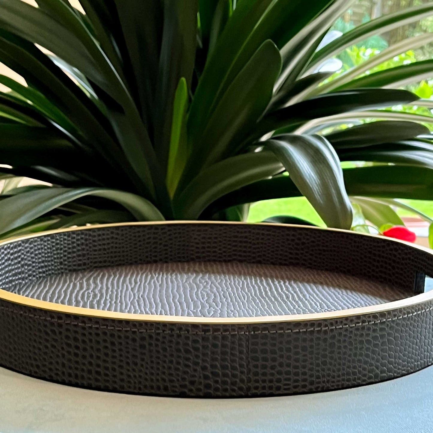 Lifestyle image view from above of round grey textured leather with gilt trim and handles on blue ottoman and green plant leaves