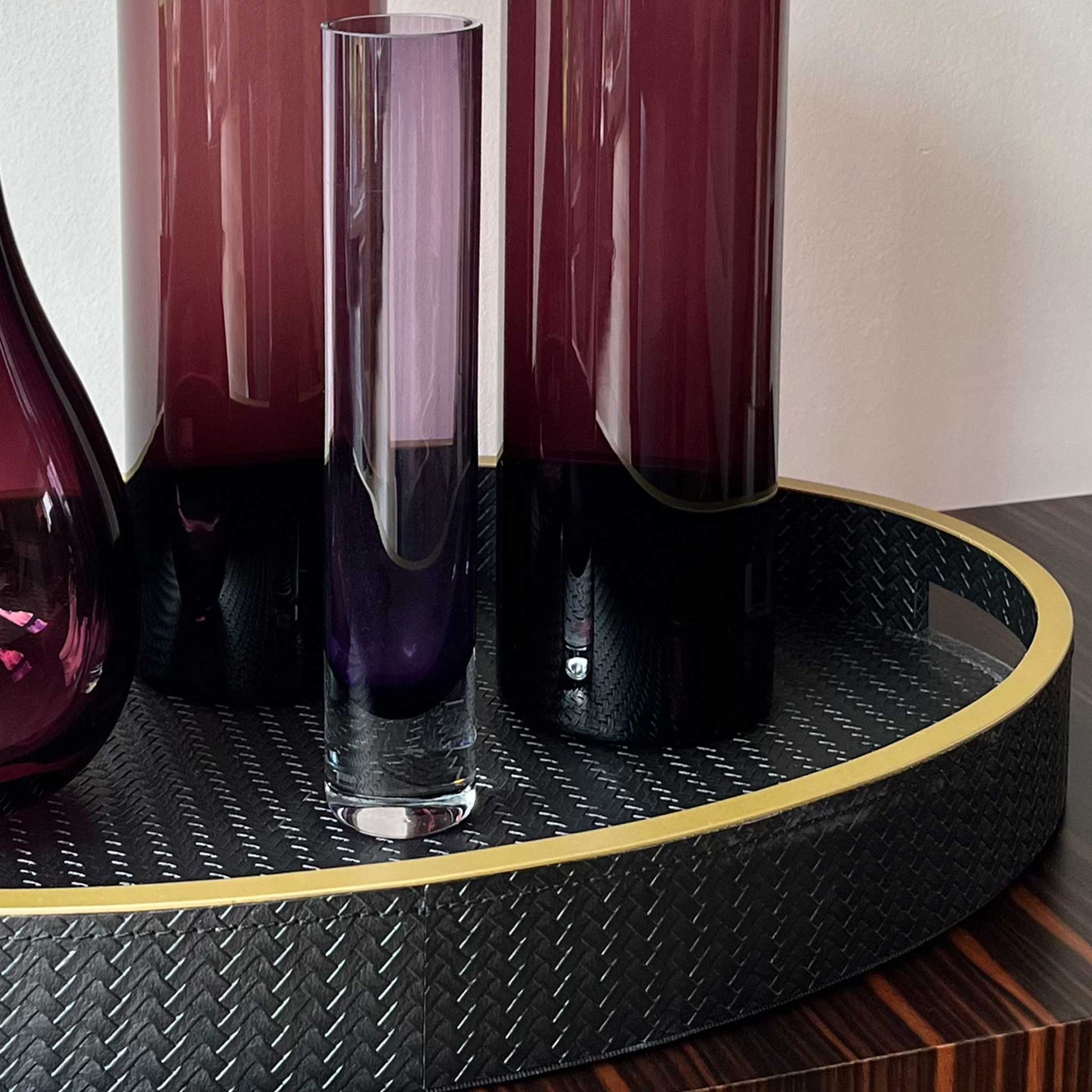 Close up of lifestyle image of oval black textured leather with gilt trim and handles. Displayed with purple vases. 