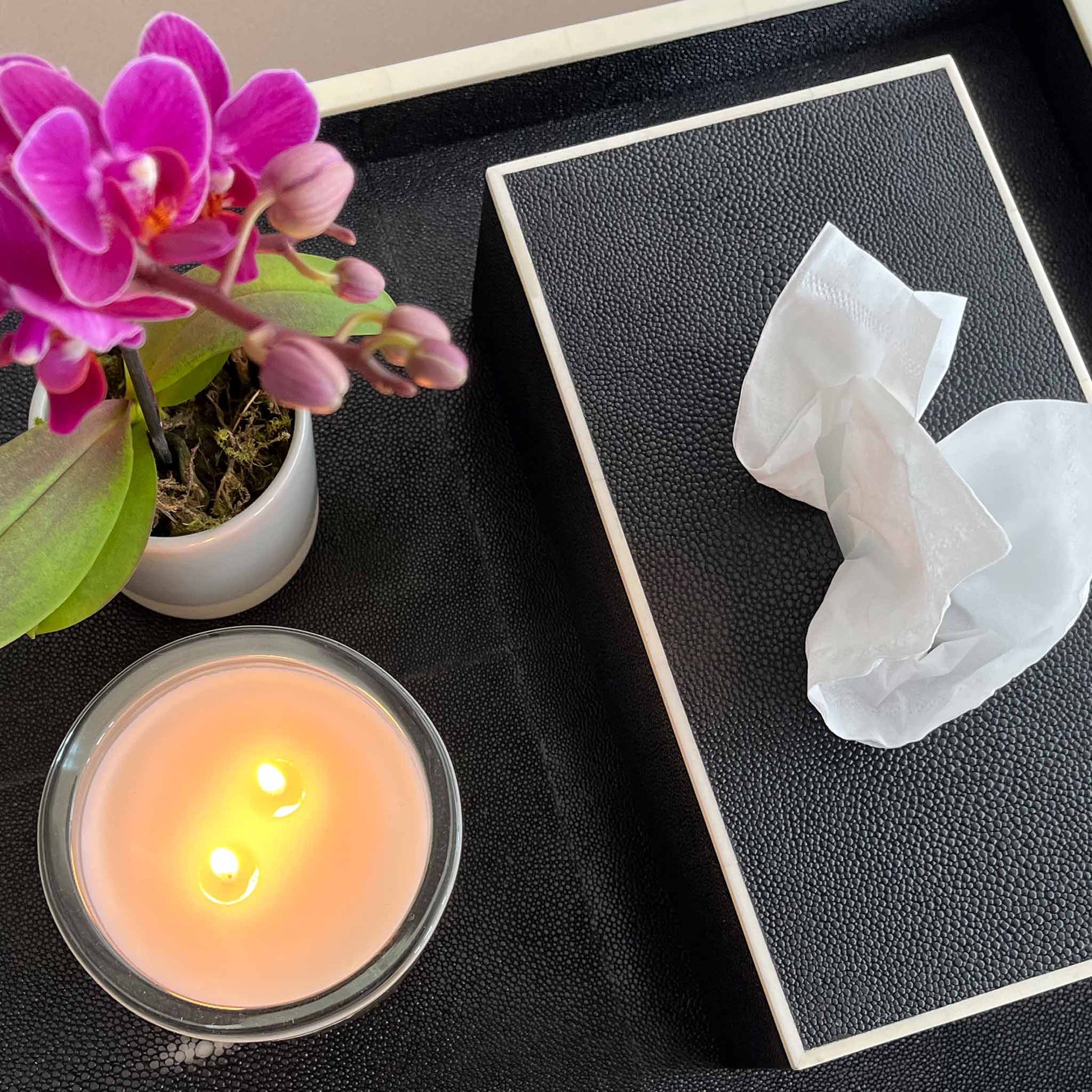 Lifestyle image of rectangular black shagreen luxury tissue box and matching tray, both with white bone trim. Displayed with lit candle and beautiful flowering plant.