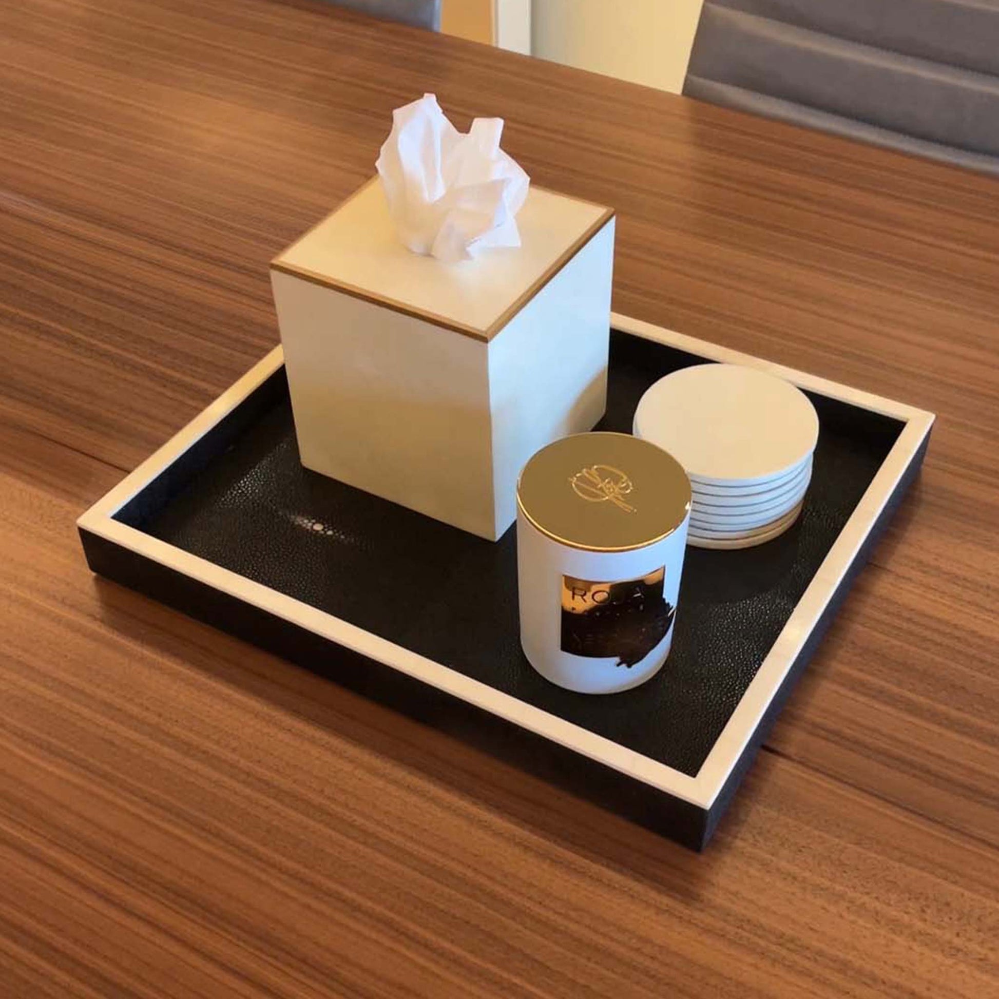 Lifestyle image of black shagreen rectangular tray with white bone trim displayed with matching parchment coasters and parchment square tissue box trimmed in brass. Displayed  in a luxury office conference table with a candle